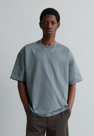 Jonge man met kort haar, dragend een loszittend grijs T-shirt, een ketting om zijn nek en donkere broek, staand tegen een eenvoudige lichte achtergrond.