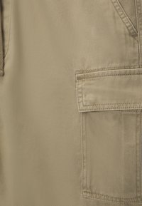 Tela beige con una textura suave que presenta un bolsillo con parche. El diseño incluye costuras visibles y un brillo sutil.