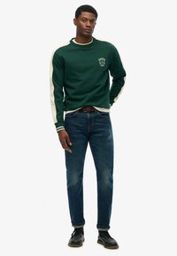 Sudadera verde con rayas blancas en los hombros, puños de canalé y cuello redondo. Combinada con jeans azul oscuros y zapatos negros.