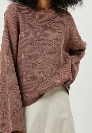 Kobieta ubrana w oversize'owy, fioletowy sweter z plecionym, ziębatym wzorem oraz kremową spódnicę, stojąca na jednolitym tle.