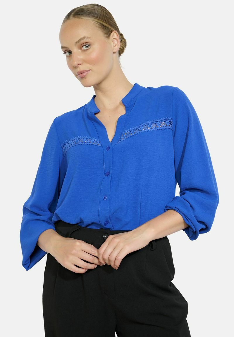 Woman wearing a blue button-up blouse with lace detail and rolled-up sleeves tucked into black high-waisted trousers, hands adjusting waistband.