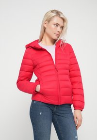 Κόκκινο μπουφάν με επένδυση και κουκούλα, κλείσιμο με φερμουάρ και ελαστικά μανίκια. Διαθέτει οριζόντιες ραμμένες ενότητες και πλαϊνές τσέπες. Φοράται με μπλε τζιν.