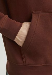 Bordowy hoodie z kieszenią kangurką; miękka, teksturowana tkanina; widoczne szwy na mankietach i u dołu; ręka spoczywa w kieszeni.