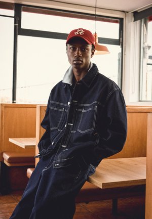 Tânăr care poartă o jachetă și pantaloni din denim închis la culoare cu cusături albe, șapcă roșie, așezat pe o bancă de lemn într-o cafenea.