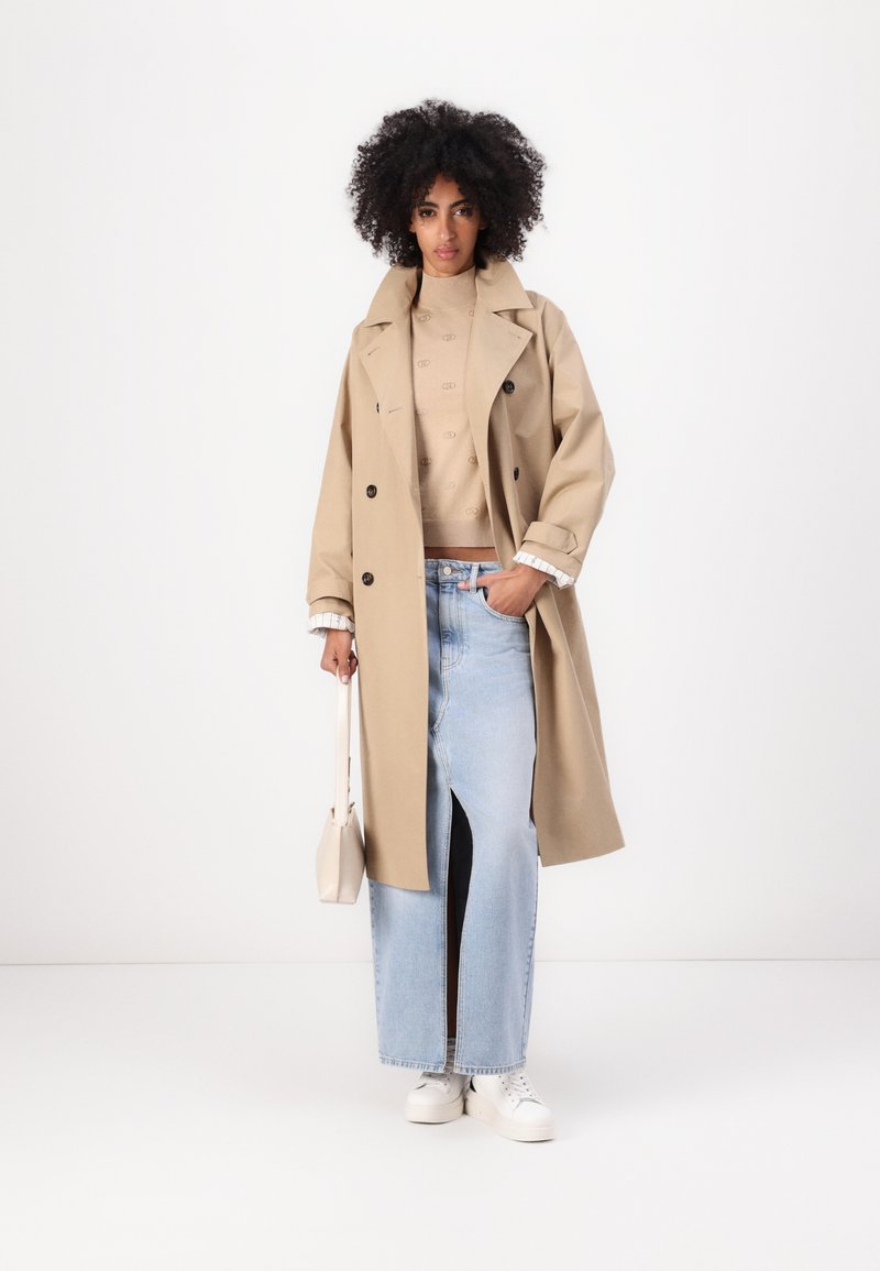 Beige Trenchcoat mit großen Knöpfen, getragen über einem hellbraunen Pullover, kombiniert mit einem Jeansrock mit hohem Sch slit und weißen Turnschuhen.
