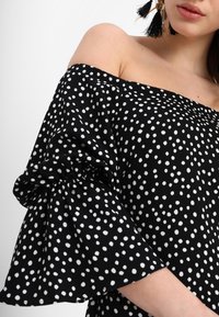 Svart off-shoulder topp med vita prickar, volangärmar och ett flödande tyg. Accentuerad med en texturerad, mjuk yta.