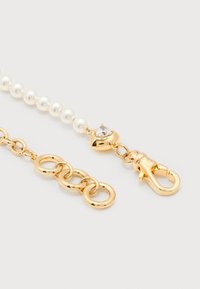 Χρυσή αλυσίδα με λευκές πέρλες, κούμπωμα σε σχήμα καρδιάς με πολύτιμο λίθο και κυκλικά κρίκια σε λευκό φόντο.