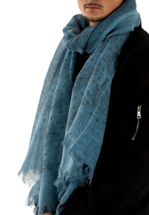 Hombre con una bufanda azul con estampado enrollada alrededor del cuello y una chaqueta negra con un bolsillo con cremallera en la manga.