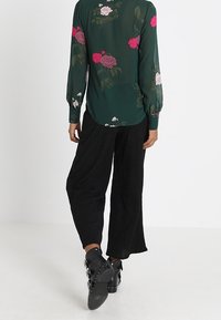 Femme portant un chemisier floral vert foncé, un pantalon noir ample et des bottines noires avec boucles, se tenant devant un fond uni.