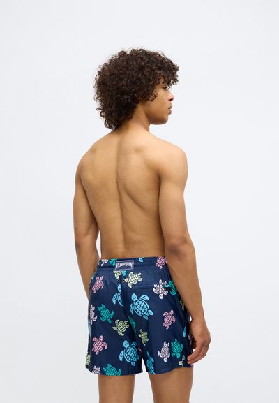 Νέος άνδρας με σγουρά μαλλιά, ντυμένος με ναυτικά μαγιό με πολύχρωμα σχέδια χελωνών, στέκεται γυμνόστηθος σε έναν απλό ανοιχτό φόντο.