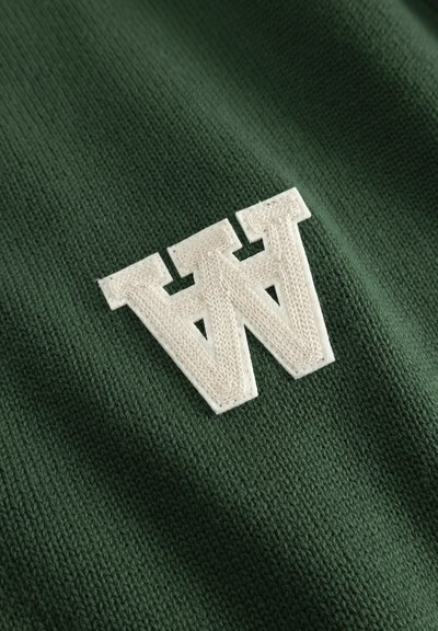 Λευκό κεντημένο έμβλημα με το γράμμα «W» σε υφή, ραμμένο σε φόντο από σκούρο πράσινο πλεκτό ύφασμα.