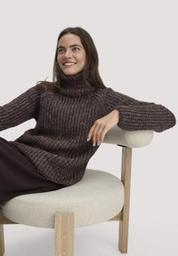 Dunkelbrauner, gestrickter Pullover mit hohem Kragen und strukturiertem Muster. Locker sitzend, kombiniert mit dunklen Hosen und einem hellen Stuhl.