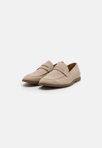 Mocassins en daim beige avec un talon bas, présentant un design cousu et deux bandes décoratives sur le cou-de-pied. Texture lisse et bout pointu.