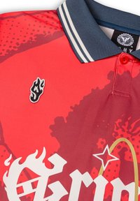 Rotes Poloshirt mit strukturiertem Design, ausgestattet mit einem marineblauen gestreiften Kragen, besticktem Flammenlogo und auffälligen weißen Grafiken mit goldenen Akzenten.