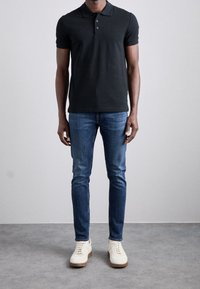 En man som bär en svart kortärmad polo, smala blå jeans och vita sneakers står på ett grått golv framför en enkel grå vägg.