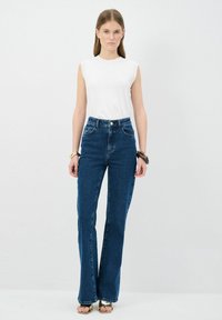 Jeans de tiro alto, de mezclilla azul oscuro, acampanados, con cinco bolsillos, cierre de botón y cremallera, combinados con una blusa blanca sin mangas ajustada.