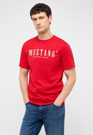 Junger Mann trägt ein rotes Mustang-T-Shirt und blaue Jeans, steht mit den Händen in den Taschen vor einem schlichten Hintergrund.