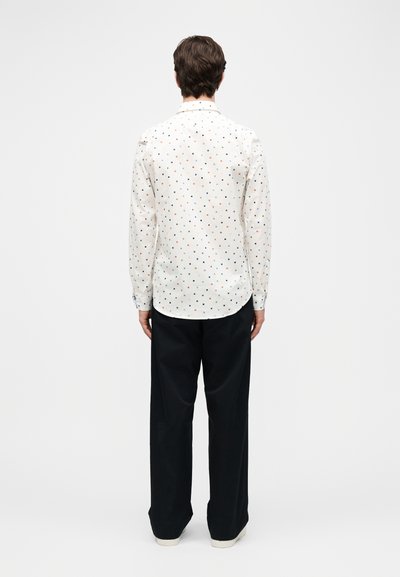 Άνδρας που φοράει λευκό μακρυμάνικο πουκάμισο με πολύχρωμα μικρά τετράγωνα, μαύρο παντελόνι και λευκά παπούτσια, στέκεται με την πλάτη σε απλό φόντο.