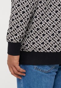 Pull en motif avec un design géométrique noir et crème, manches longues et ourlet côtelé noir, associé à un jean bleu clair.
