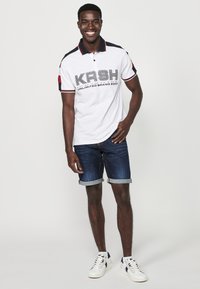 Bijela polo majica s mornarsko plavim i crvenim detaljima, s grafičkim logotipom. Kombinira se s preklopljenim trapericama i bijelim tenisicama.