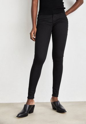 Personne portant un pantalon noir cintré, un haut noir sans manches et des chaussures noires à talons sans lacets, debout sur un sol clair.