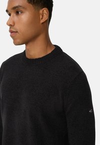 Junger Mann in einem schwarzen Strickpullover mit Rundhalsausschnitt, der nach links vor einem schlichten hellen Hintergrund schaut.