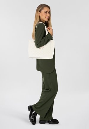 Borsa tote bianca trapuntata con due manici superiori, caratterizzata da accenti metallici. Il modello indossa un abito verde con pantaloni larghi e scarpe nere.