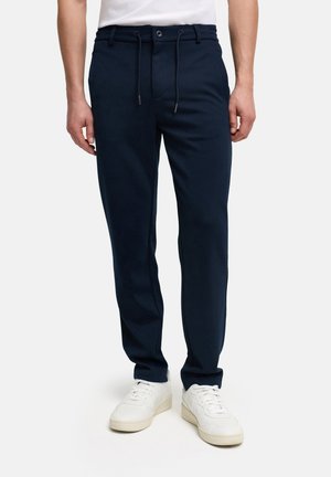 Mand iført marineblå bukser med snøre og hvide sneakers, stående mod en ensfarvet hvid baggrund.