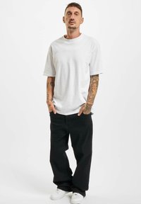 Weißes Baumwoll-T-Shirt mit kurzen Ärmeln, kombiniert mit schwarzen weit geschnittenen Hosen und weißen Sneakers. Tattoos sind an den Armen sichtbar.