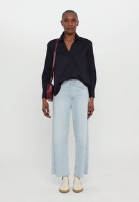 Svart långärmad skjorta, ljusblå vida jeans med sidöppningar och krämfärgade sneakers. Liten vinröd crossbody-väska. Släta tygtexturer.