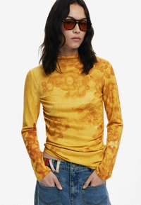 Langärmliges gelbes Oberteil mit einem strukturierten Blumenmuster. Der Stoff hat eine gerippte Oberfläche und ist mit blauen Jeans kombiniert, die Hände in den Taschen.
