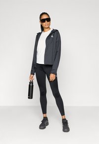 Ελαφρύ, σκούρο γκρι μπουφάν με κουκούλα, λευκό μπλουζάκι, μαύρο κολάν και μαύρα αθλητικά παπούτσια. Κρατάει μια μαύρη παγούρι νερού. Γυαλιά ηλίου.