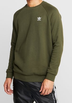 Homme portant un sweat-shirt Adidas vert olive avec logo sur la poitrine et un pantalon noir avec une bande blanche sur le côté et du texte.