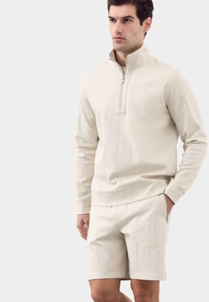 Uomo che indossa un top crema a maniche lunghe con zip e pantaloncini abbinati, in piedi con una mano nella tasca dei pantaloncini, guarda di lato.