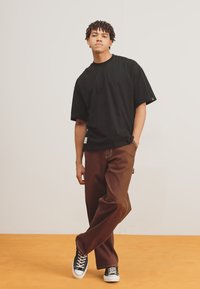 Sort kortærmet t-shirt parret med brune brede bukser. Outfittet har hvid syning og sorte sneakers. Stående mod en ensfarvet baggrund.