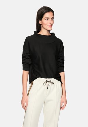 Black pullover with a high, folded collar and long sleeves, made of soft fabric. Paired with white pants featuring side stripes and drawstring.