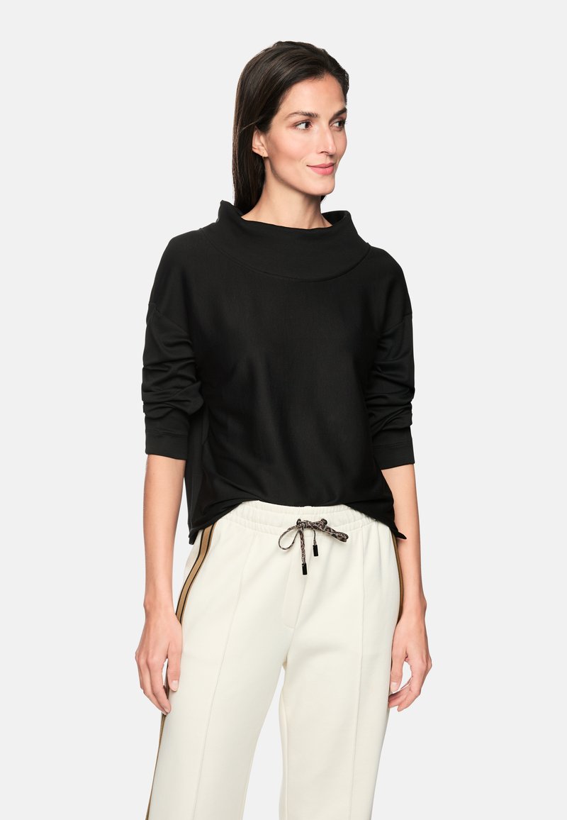 Schwarzer Pullover mit hohem, umgeschlagenem Kragen und langen Ärmeln, aus weichem Stoff. Kombiniert mit weißen Hosen mit seitlichen Streifen und Kordelzug.