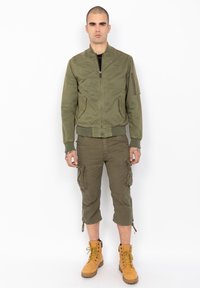 Chaqueta bomber verde olivo con cremallera en la parte delantera, puños acanalados y bolsillos laterales, combinada con pantalones cortos cargo de longitud hasta la rodilla y botas de trabajo color marrón claro.