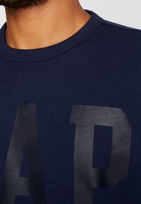 Navy modrý svetřík s kulatým výstřihem, na přední straně s velkými černými tištěnými písmeny. Měkký materiál z bavlněné směsi s hladkým povrchem.