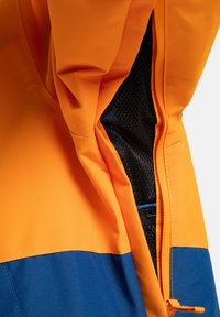 Zbliżenie na pomarańczową i niebieską kurtkę z częściowo rozpiętym panelem wentylacyjnym z siatki po boku.