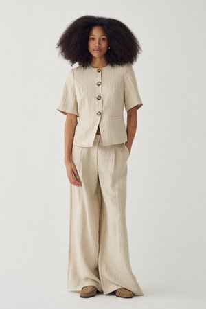 Naine mahuka lokkis juustega, kes kannab beežikat lühikeste varrukatega nööpidega jopet ja laia säärega pükse pruuni mokassiinidega.