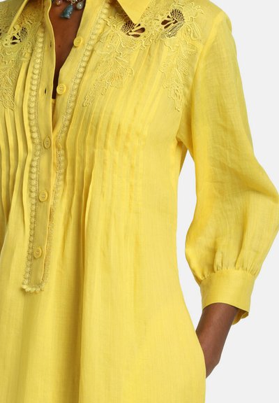 Žltá blúzka s dlhým rukávom, skladaná s kvetinovou čipkovou výšivkou na ramenách, predným zapínaním na gombíky a manžetovými rukávmi.