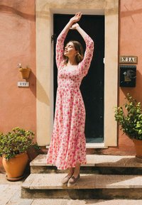 Roze bloemen midi-jurk met lange mouwen en gerimpelde corsage, met een gelaagde rok. Model staat op stenen trappen met groenvoorziening in de nabijheid.