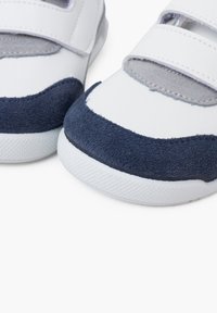 Baskets blanches avec des accents en suède bleu marine et des détails en textile gris. Fixées avec deux bandes Velcro. Semelle en caoutchouc texturé.