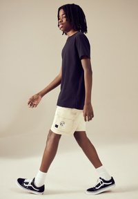 Black cotton t-shirt, beige cargo shorts with embroidered design, and black shoes with white laces. Simple, casual outfit in relaxed fit.