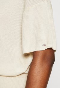 Manche de pull en tricot beige avec une étiquette de logo sur le poignet, associée à un bras de peau foncée, mettant en valeur la texture et le contraste des couleurs.