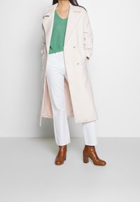 Femme portant un long manteau crème, un pull en tricot vert, un pantalon blanc et des bottines à talons marron, se tenant devant un fond blanc uni.