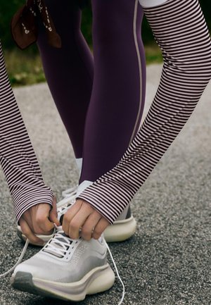 Handen die witte hardloopschoenen strikken bij een persoon die gestreepte lange mouwen en paarse leggings draagt op een buitenpad.