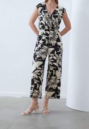 Vrouw die een zwart-beige jumpsuit met wijde pijpen en bloemenprint draagt, met kapmouwen en witte sandalen met blokhakken en smalle bandjes, staand binnen.