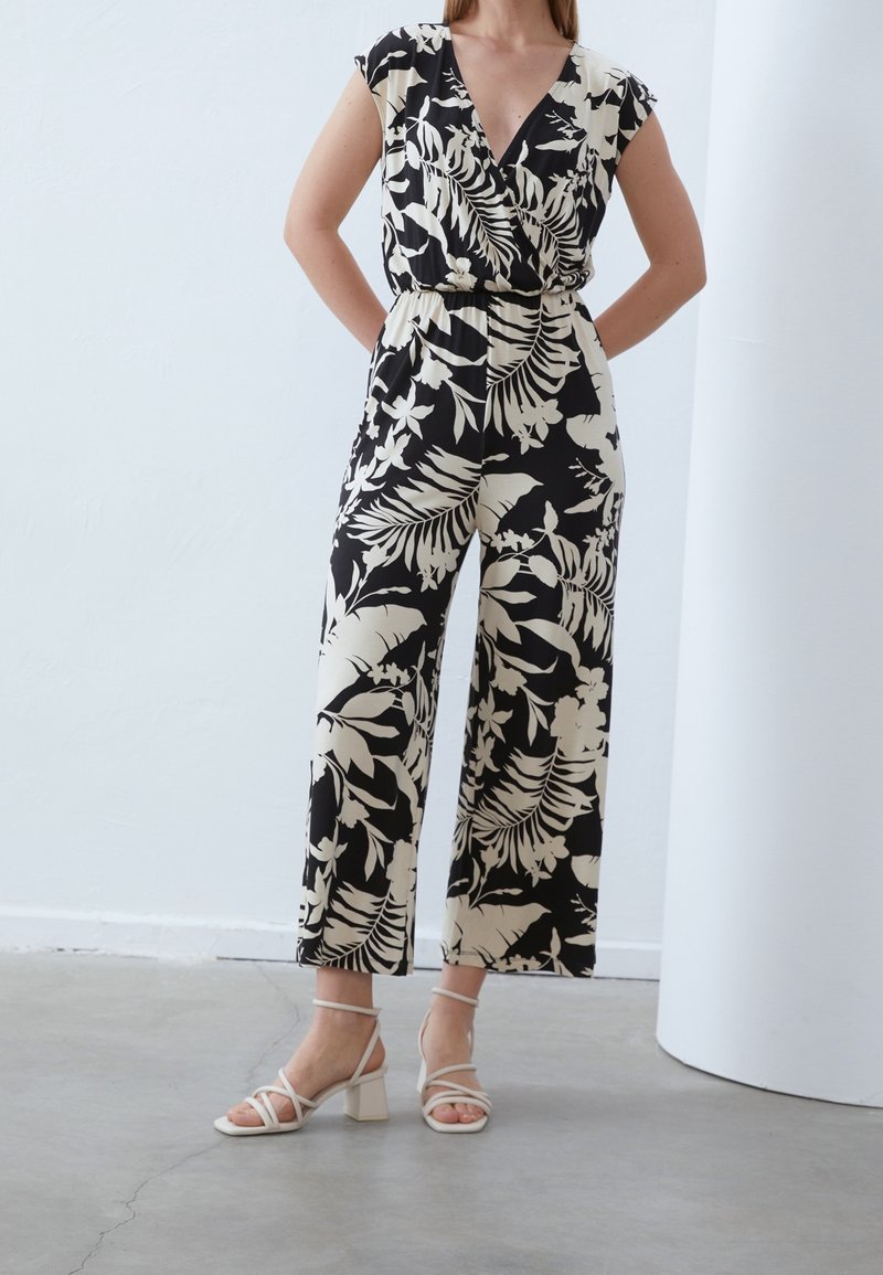Femme portant une combinaison pantalon ample à fleurs noires et beiges avec des manches courtes à cape et des sandales blanches à talons blocs et brides, debout à l'intérieur.